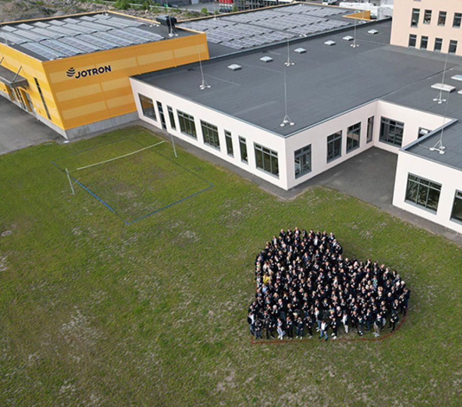 Jotron employees stading in a heart-shaped group in front of the Jotron premises. Solar-panels on the building's roof.