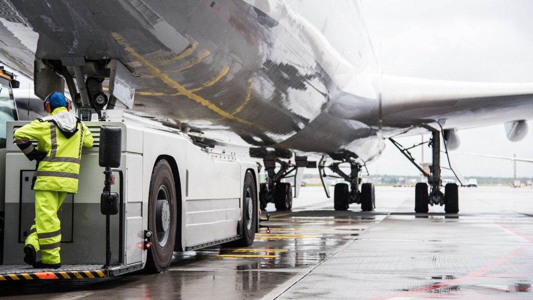 Large plane at airport during pushback with ground crew person in yellow clothes standing on vehicle.